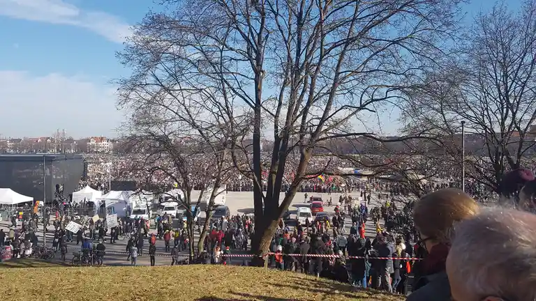 A wide-angle photo shows a massive crowd gathered in a public square for what appears to be a protest or rally, with bare…