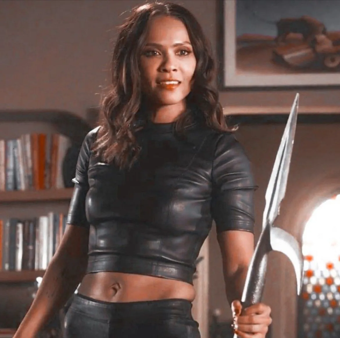 A woman with long dark hair, wearing a black leather crop top and pants, holds a large silver weapon while smiling…