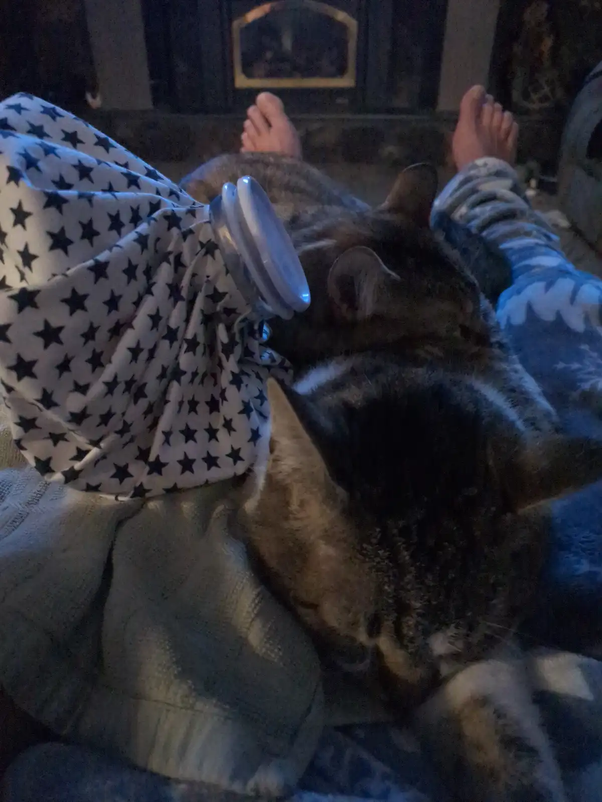 A person in pajamas lies on a couch with their feet up, a tabby cat curled up on their lap, and a small white fan resting on…