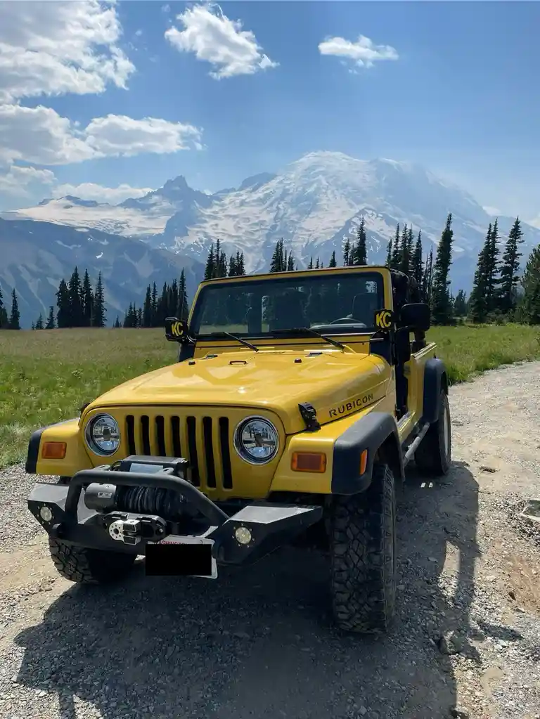 A bright yellow Jeep Wrangler Rubicon is parked on a gravel road in front of a majestic snow-capped mountain, likely Mount…