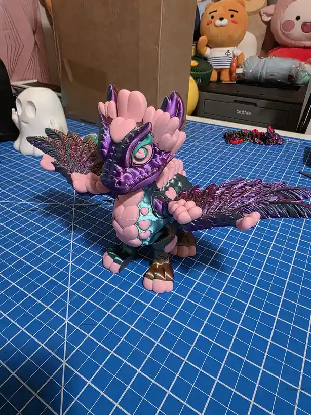 A custom-painted, stylized dragon figurine with pink, purple, and teal metallic colors sits on a blue grid cutting mat. The…