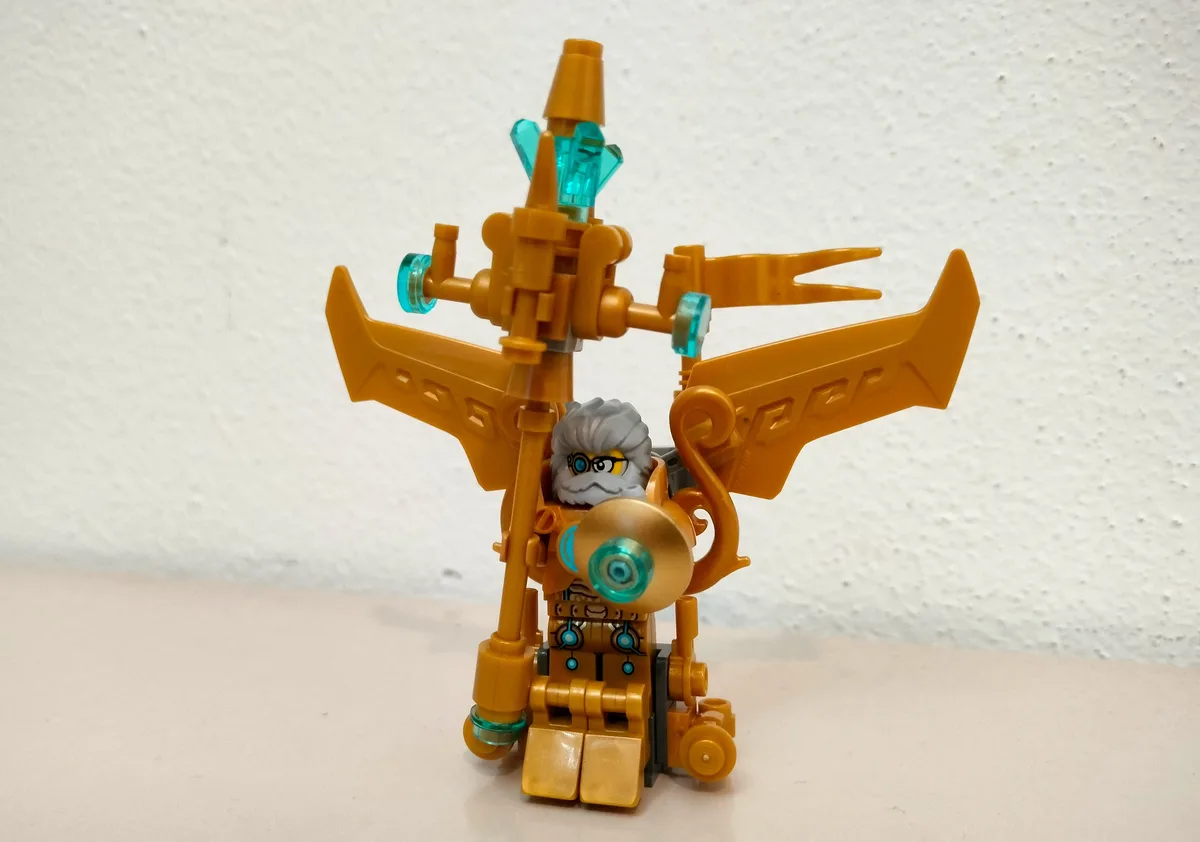 A LEGO minifigure with gray hair and glasses is seated in a complex, golden mechanical contraption with large wing-like…