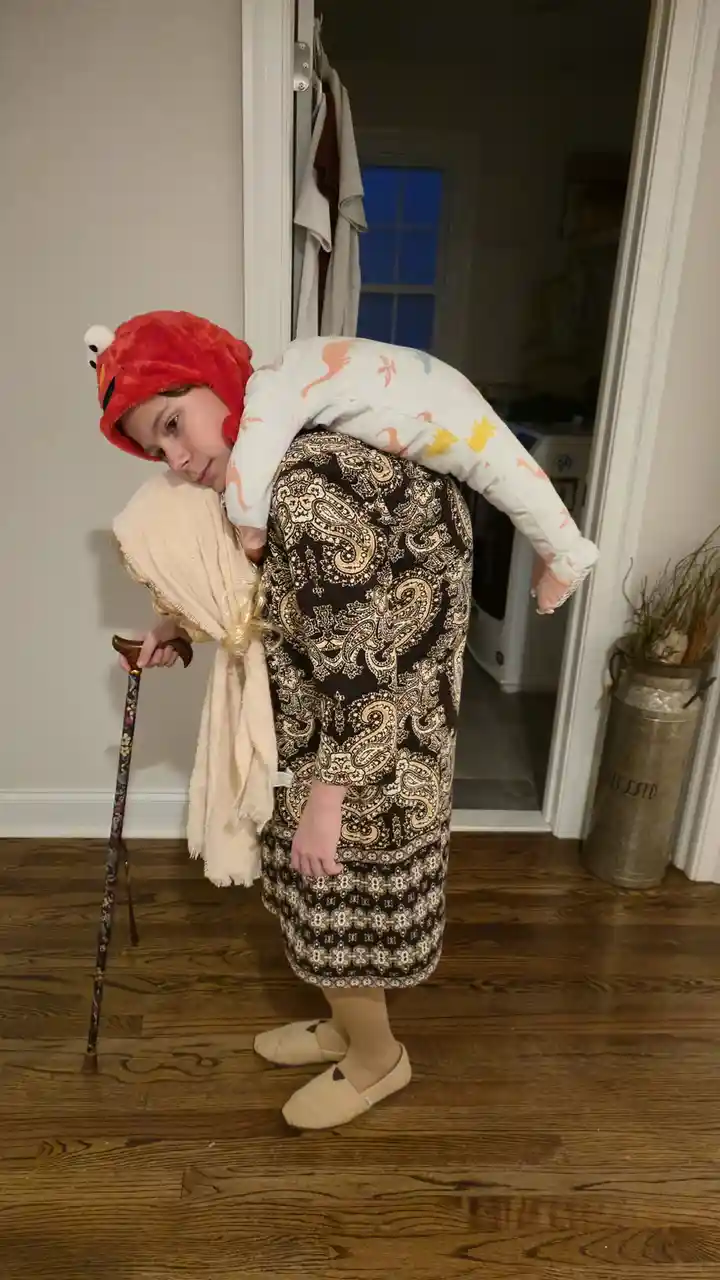 A person dressed as an elderly woman with a cane and headscarf is bent over, supporting another person wearing a red Elmo…
