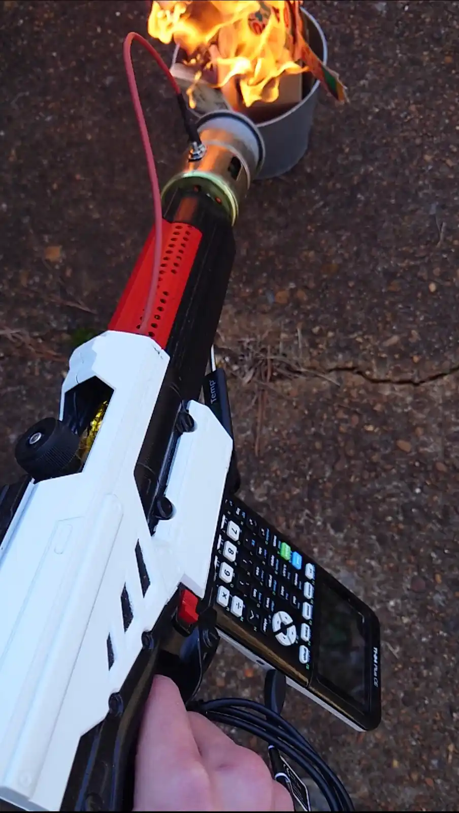A hand holds a white and black toy gun modified to look like a flamethrower, with a red hose connected to a metal nozzle…