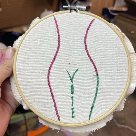 A hand holds a wooden embroidery hoop with white fabric stretched taut. Embroidered in pink thread are two curved lines…
