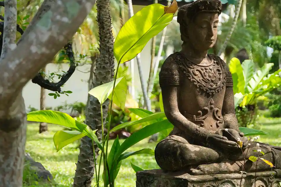 A serene stone statue of a person in a meditative pose sits in a lush, tropical garden with large green leaves and trees…