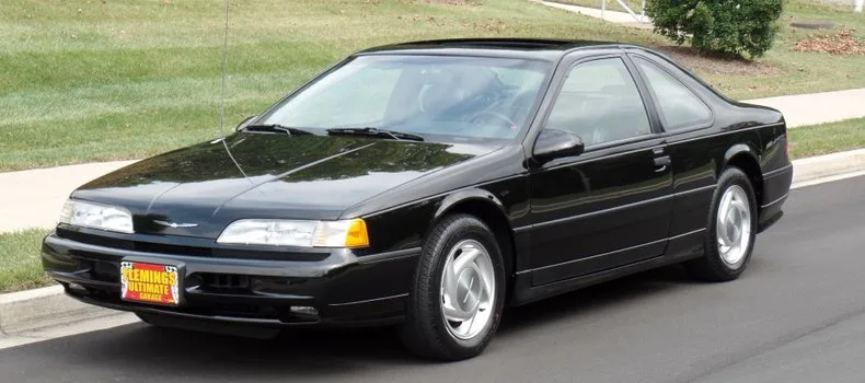 A black 1990s Ford Taurus coupe is parked on a street with a grassy area behind it. The car has a custom license plate that…