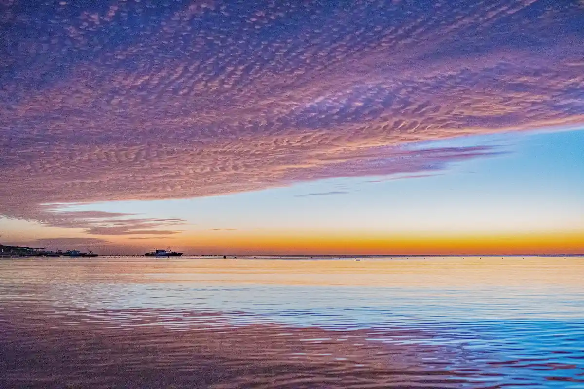 A serene sunset over water with vibrant purple, pink, and orange clouds reflecting on calm ripples. Silhouettes of boats and…