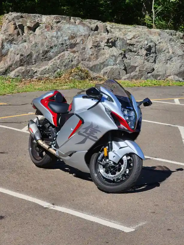 A silver and red Suzuki sport motorcycle is parked in a marked parking space on asphalt, with a large rocky cliff face and…