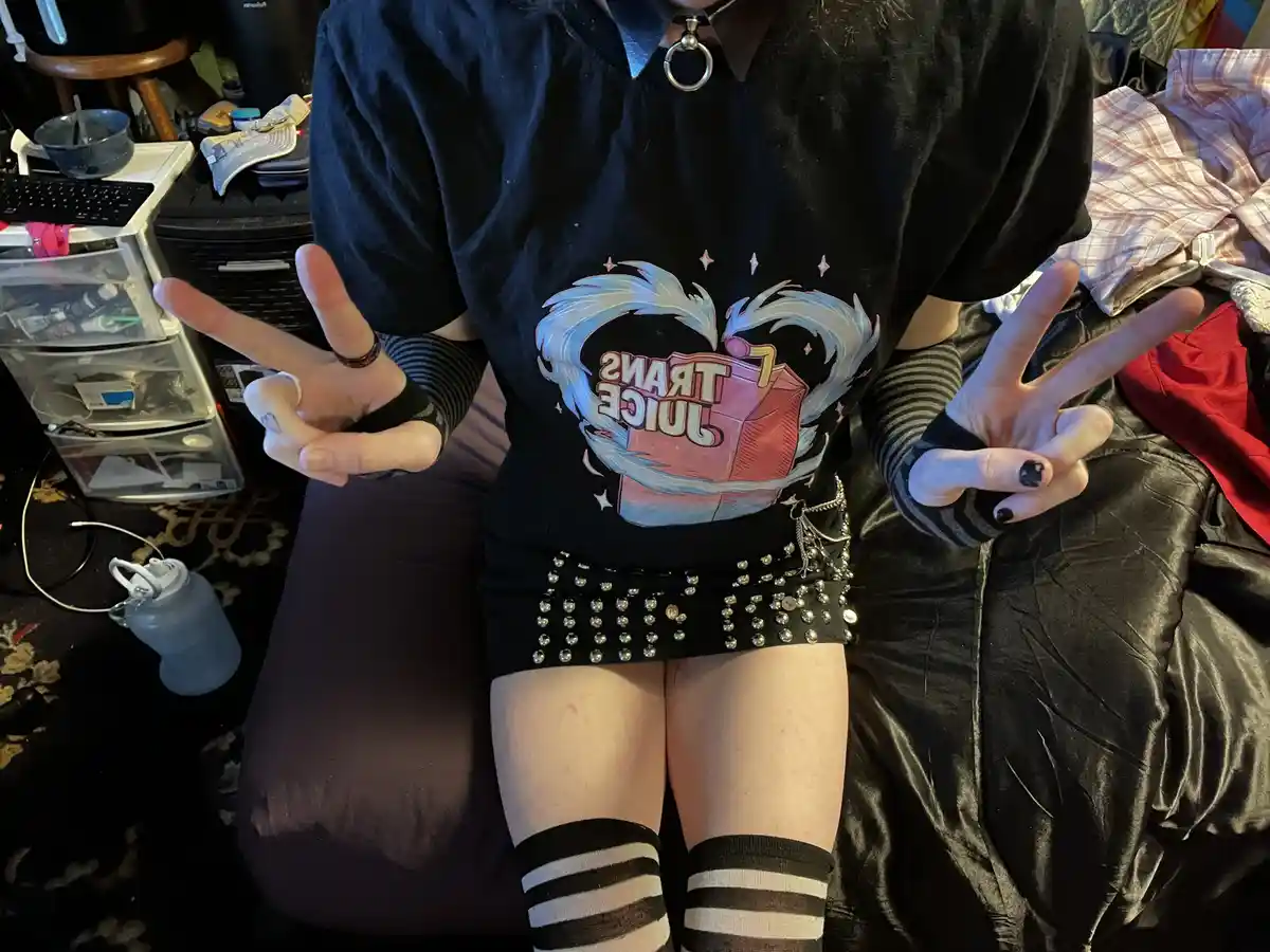 A person is sitting on a bed in a messy room, wearing a black t-shirt with a graphic of a juice box inside a heart and the…