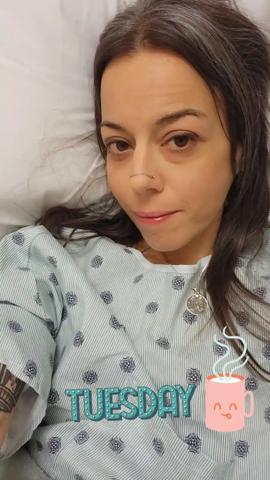 A woman with dark hair and a nose ring lies in bed wearing a patterned hospital gown, looking tired and unenthusiastic…