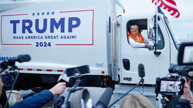A man wearing an orange safety vest sits in the driver's seat of a white semi-truck, smiling and waving. The truck's side…