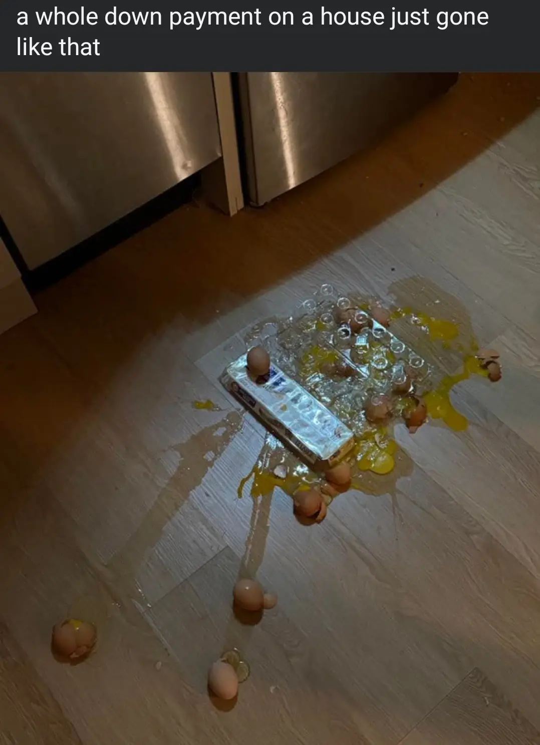 A spilled egg carton lies on a light wood floor, with broken eggs, yolks, and shells scattered around it, next to stainless…
