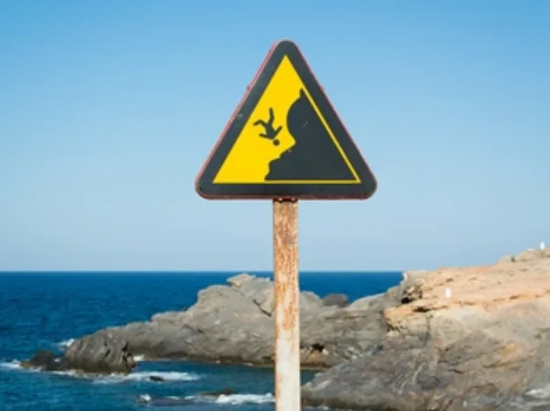 A yellow triangular warning sign with a black silhouette of a person falling off a cliff is mounted on a rusty pole near a…