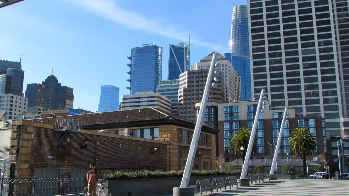 A man in an orange jacket stands beside a bicycle in a city plaza with modern skyscrapers in the background. The image has…