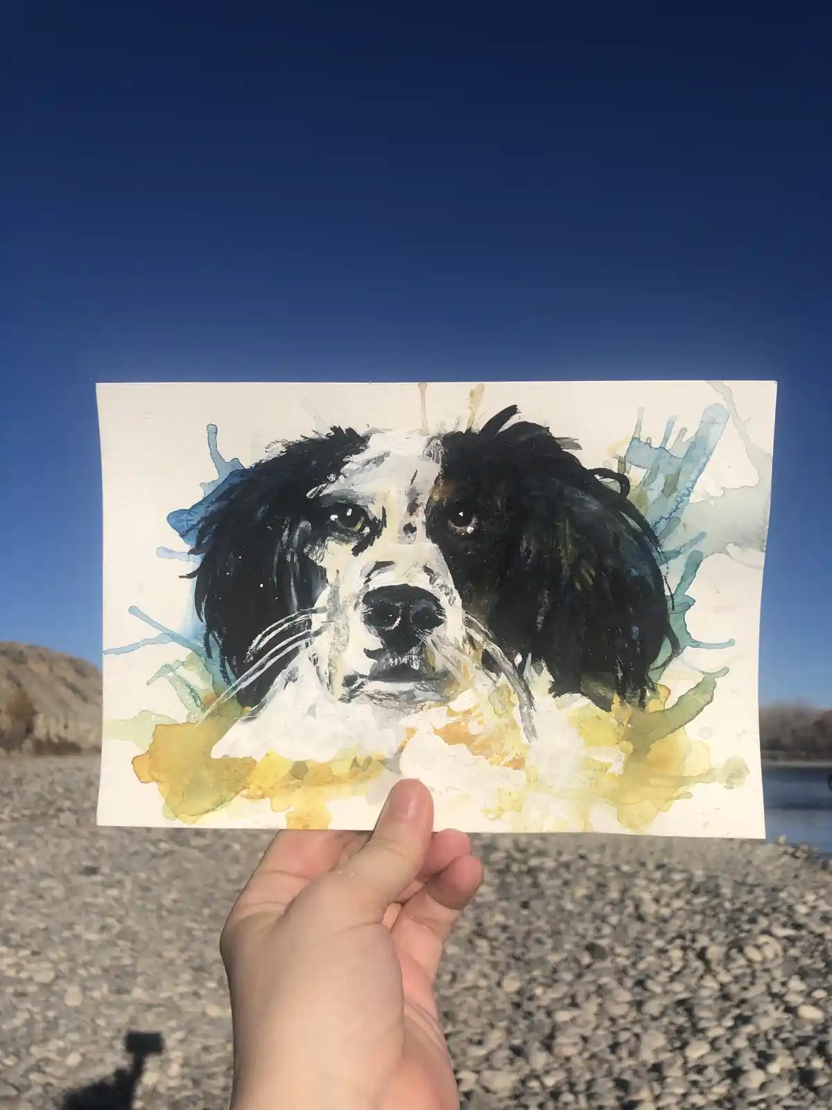 A hand holds up a watercolor painting of a black and white dog with expressive eyes, set against a backdrop of a clear blue…