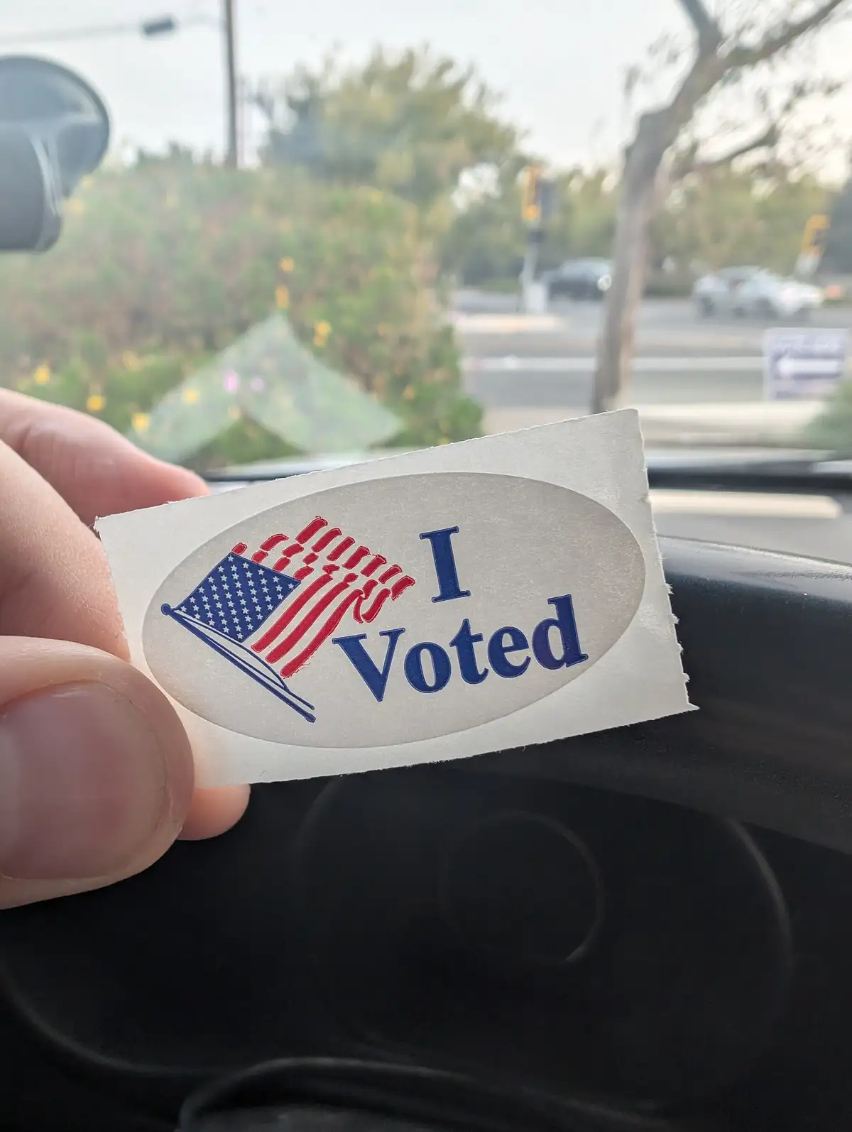 A hand holds up a standard 'I Voted' sticker featuring an American flag design inside an oval, taken from inside a car with…