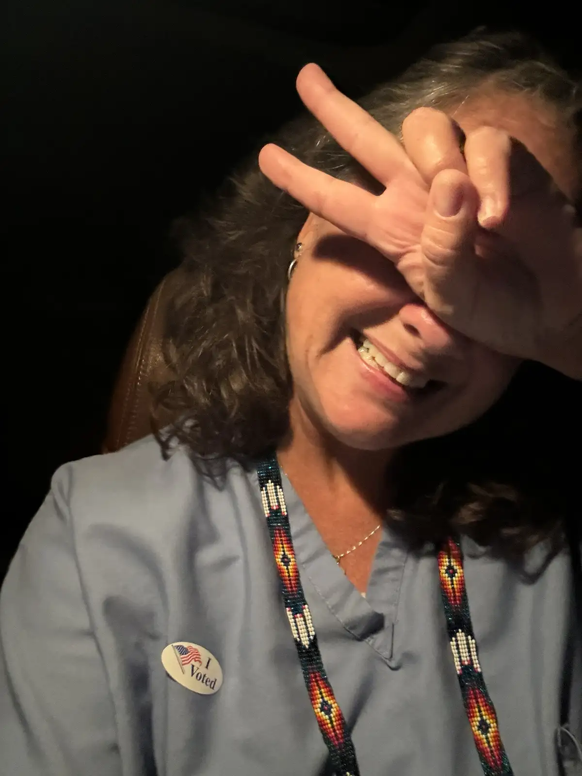 A smiling woman with gray hair, wearing a light blue shirt, covers her eyes with her hand while making a peace sign. She has…