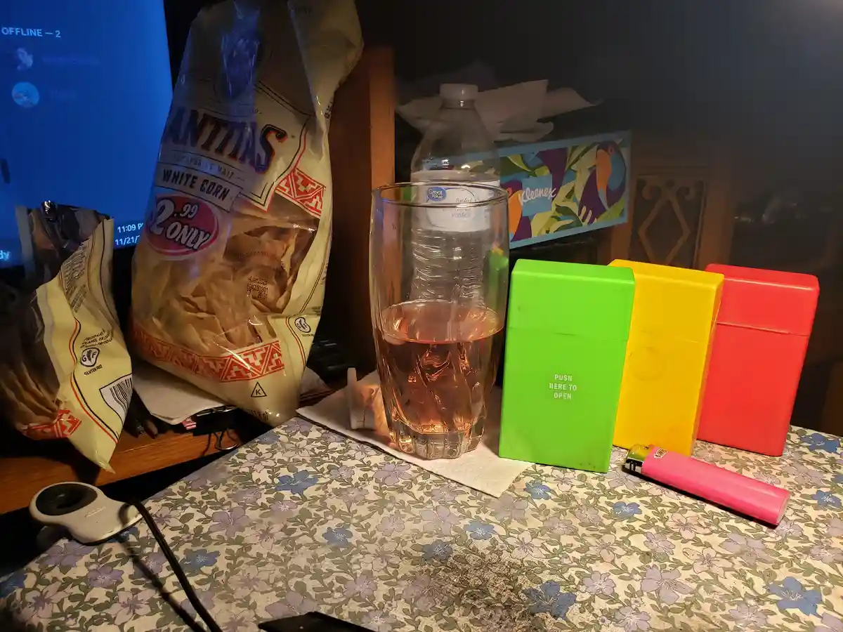 A cluttered table holds a bag of Frito-Lay tortilla chips, a glass of pink liquid, a water bottle, and three colorful…
