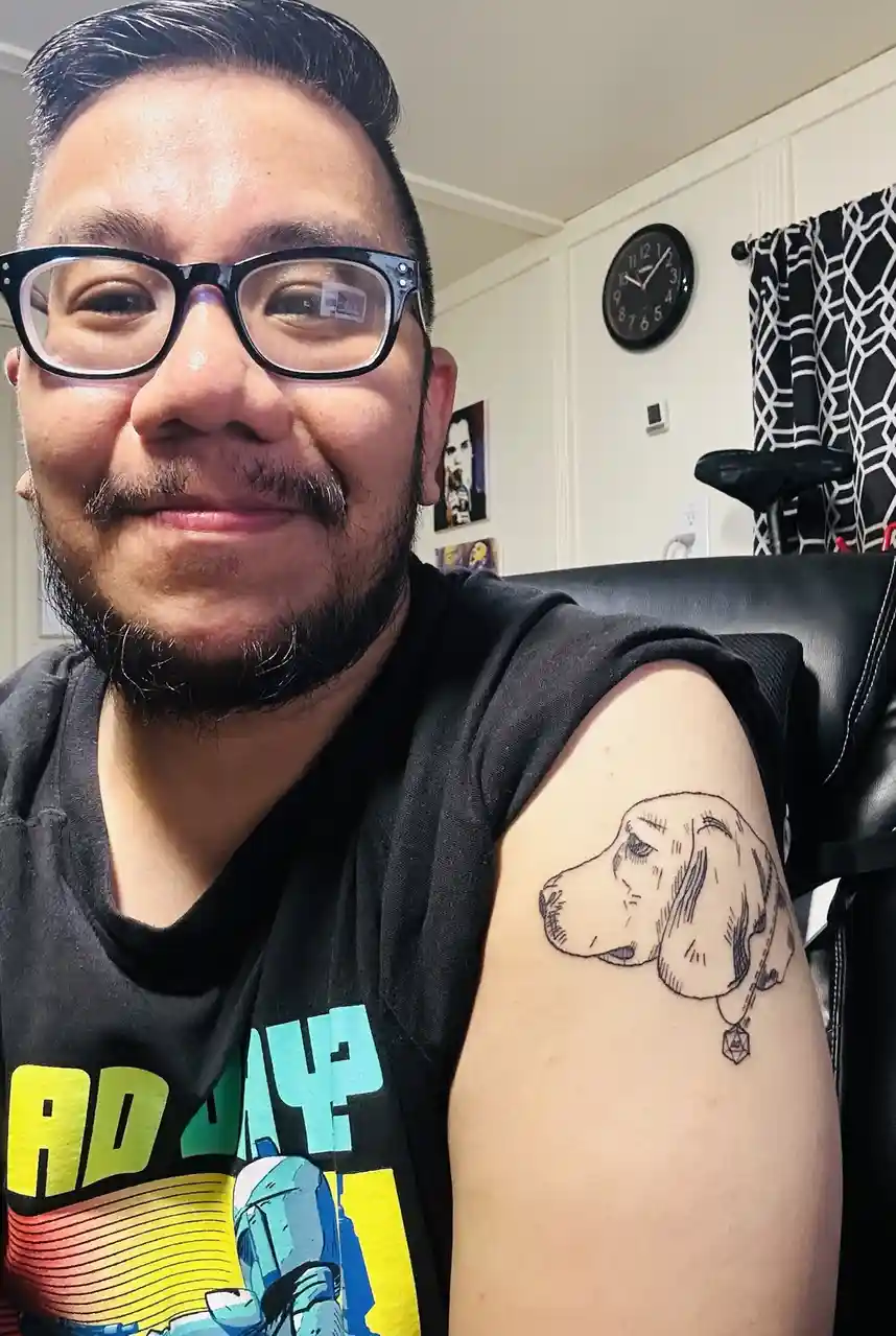 A man with glasses and a beard smiles while showing off a black-and-white tattoo of a dog's head on his upper arm. The…