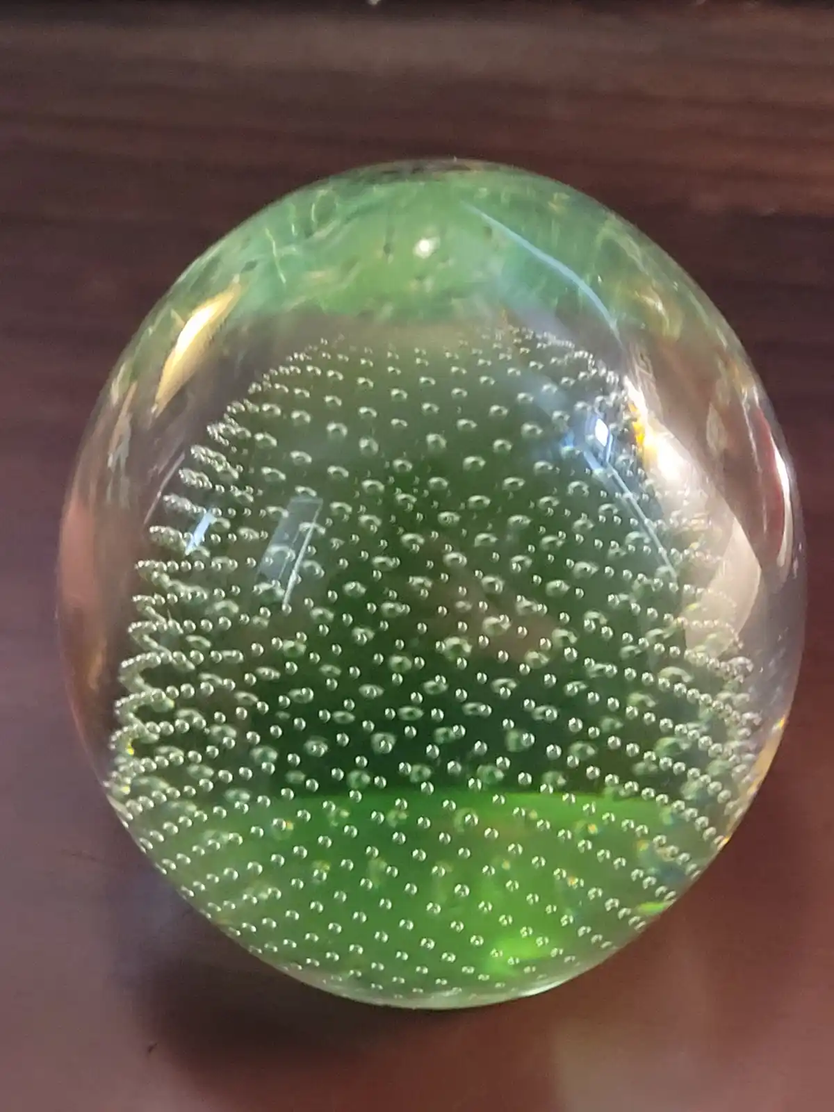 A close-up photo shows a green glass paperweight with a textured, bubble-like interior pattern, resting on a wooden surface…