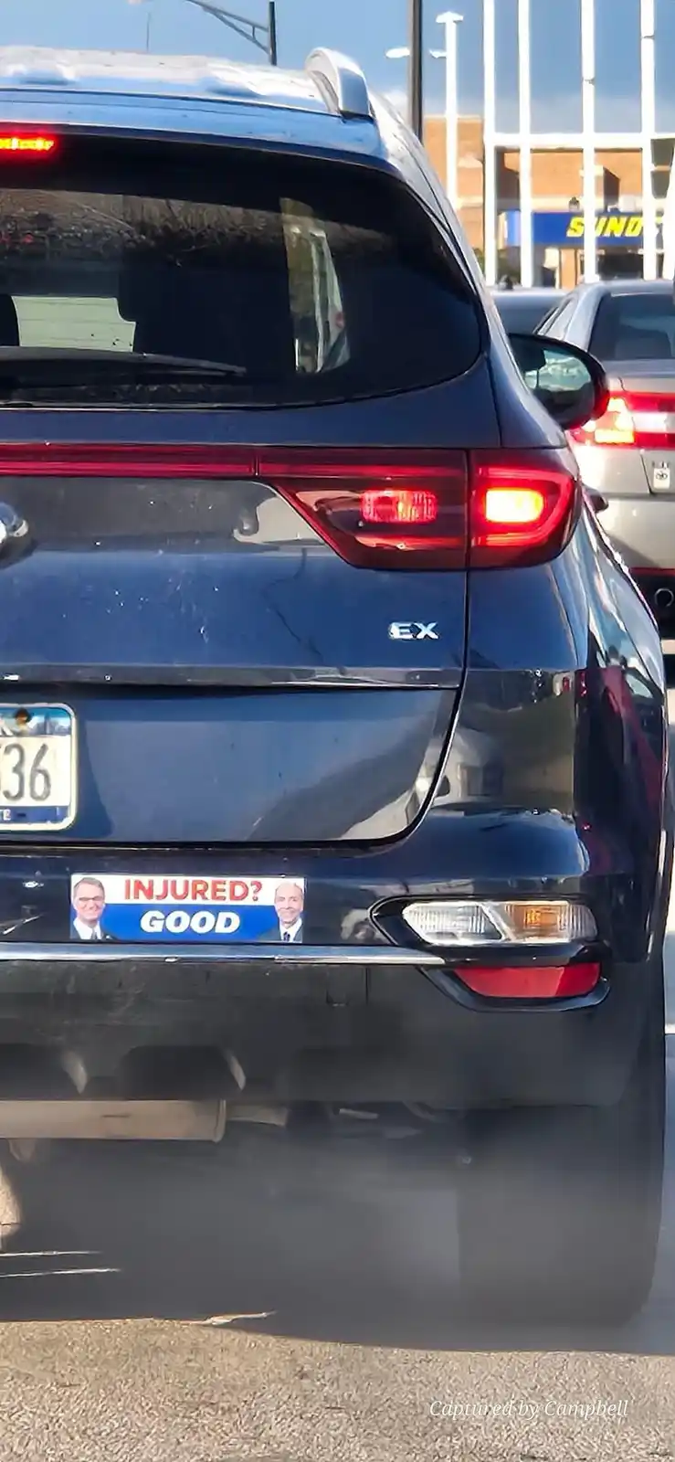 The rear of a dark blue SUV is shown in traffic, featuring a bumper sticker with two smiling men and the text 'INJURED?…