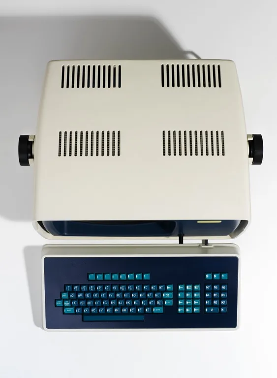 A top-down photo of a beige, vintage, portable computer with a dark blue keyboard featuring light blue keys. The machine has…