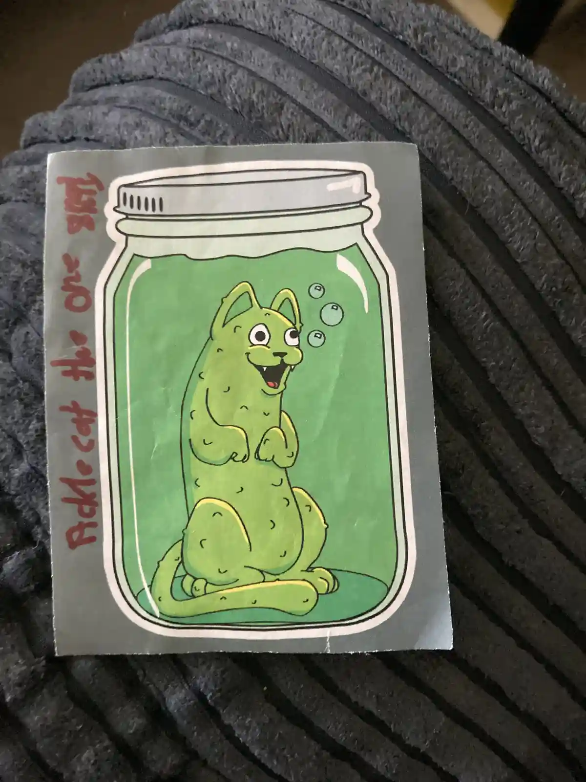 A sticker depicts a cartoon cat with a pickle-like green body, sitting inside a glass jar filled with green brine and…