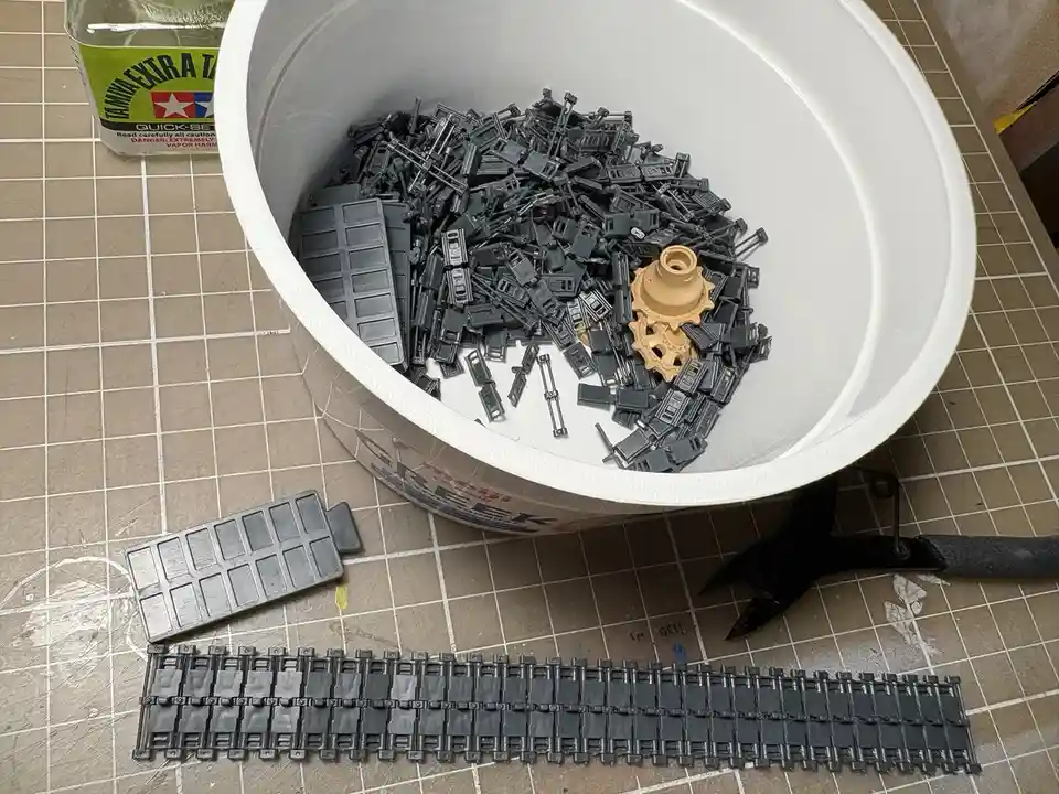 A white bucket filled with numerous small, gray plastic model parts sits on a grid-patterned work surface, with a single tan…