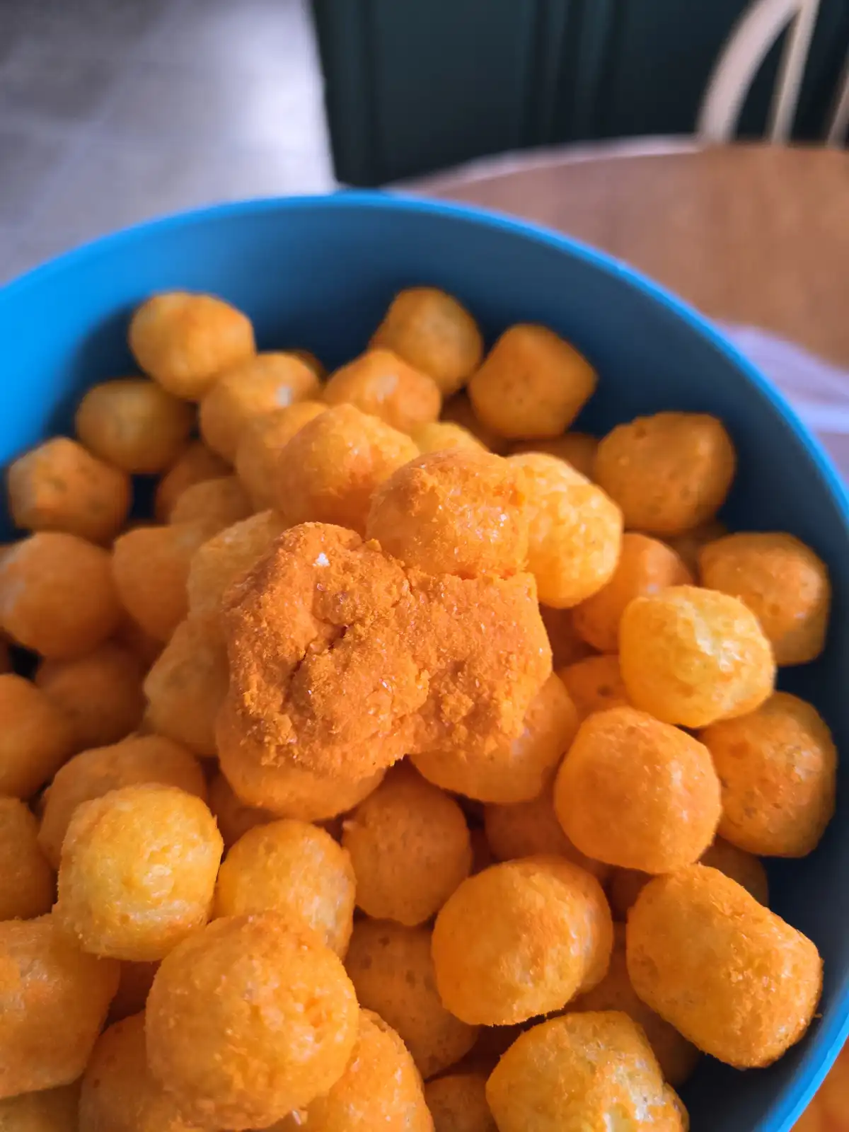 A close-up photo shows a blue bowl filled with orange, round, cheese-flavored snack balls, with one particularly large…