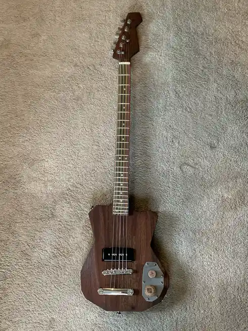 a dark wood electric guitar with a unique body shape lies on a beige carpet. The guitar has a standard neck and headstock…