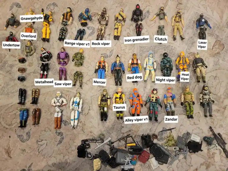 An overhead shot displays numerous G.I. Joe action figures arranged in rows on a patterned carpet, each labeled with a name…