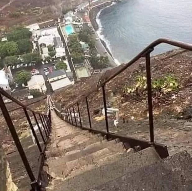 a steep, weathered concrete staircase with metal railings descends a rocky hillside overlooking a coastal town and ocean…
