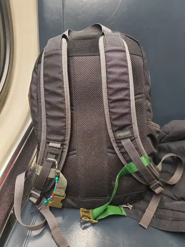 A dark gray Timbuk2 backpack sits on a blue vinyl train seat, its straps and buckles visible. A green strap and a beaded…