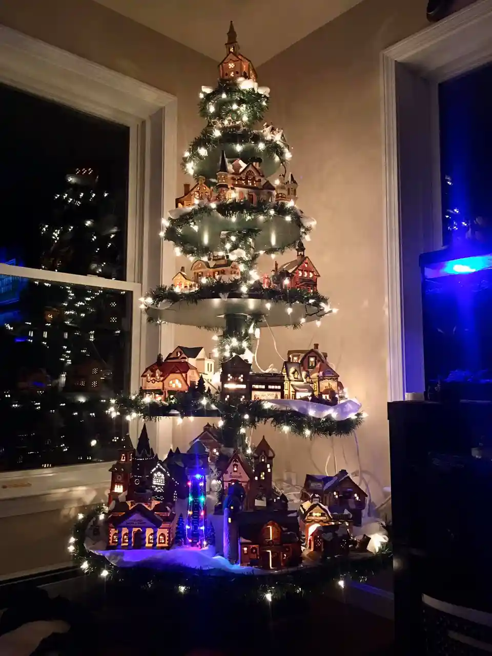 A multi-tiered, artificial Christmas tree decorated with miniature, illuminated buildings resembling a complete village…