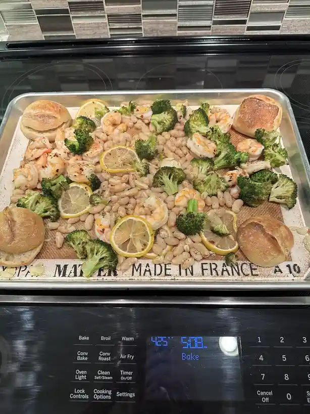 A metal baking tray holds a large, messy assortment of roasted shrimp, broccoli, white beans, lemon slices, and small bread…