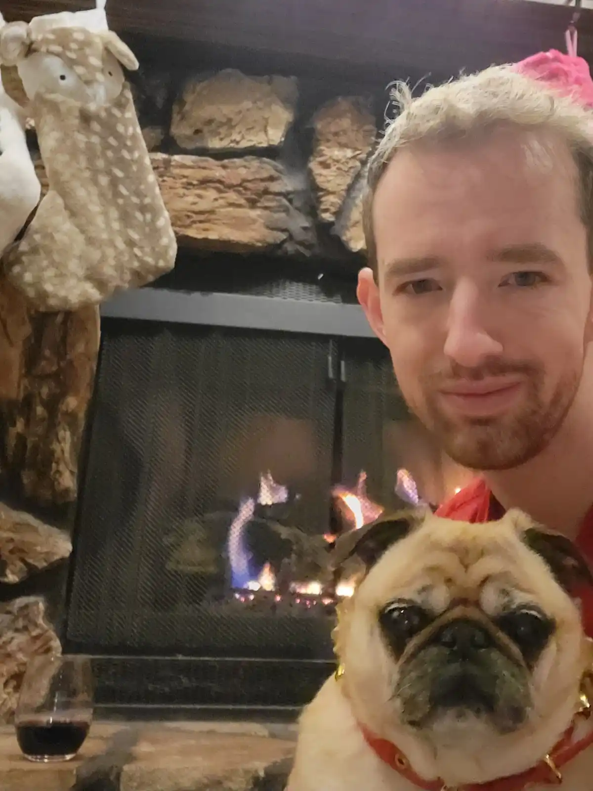 A man with blonde hair and a slight smile poses with a pug dog in front of a lit fireplace. The pug, wearing a red collar…