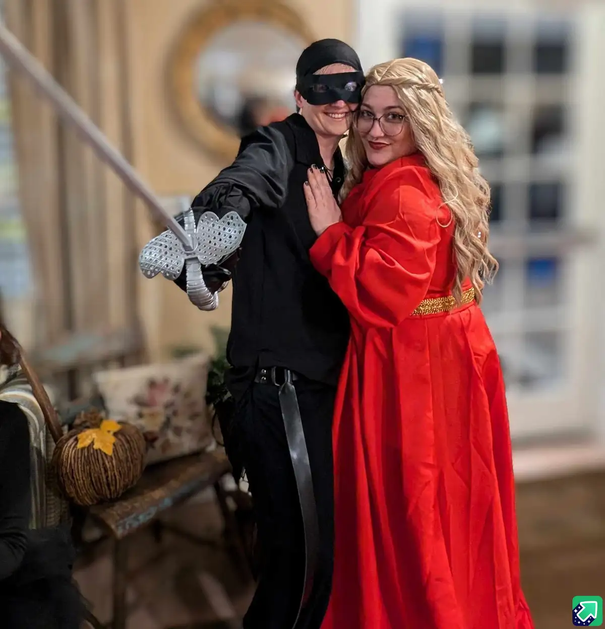 Two women pose together indoors; one wears a black costume with a blindfold and holds a sword, while the other wears a red…