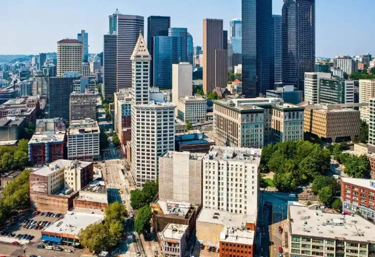 An aerial photo of a dense urban skyline with numerous skyscrapers and lower buildings under a clear blue sky. Overlaid text…