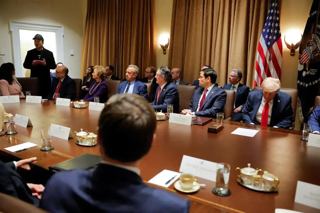 A photo shows a formal meeting in a large room with a long wooden table, American flags, and officials seated. A man in a…