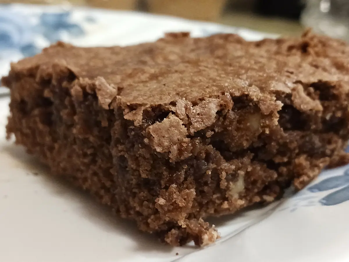 A close-up photo shows a square, dense, crumbly brownie-like dessert on a white plate with blue floral patterns. The dessert…