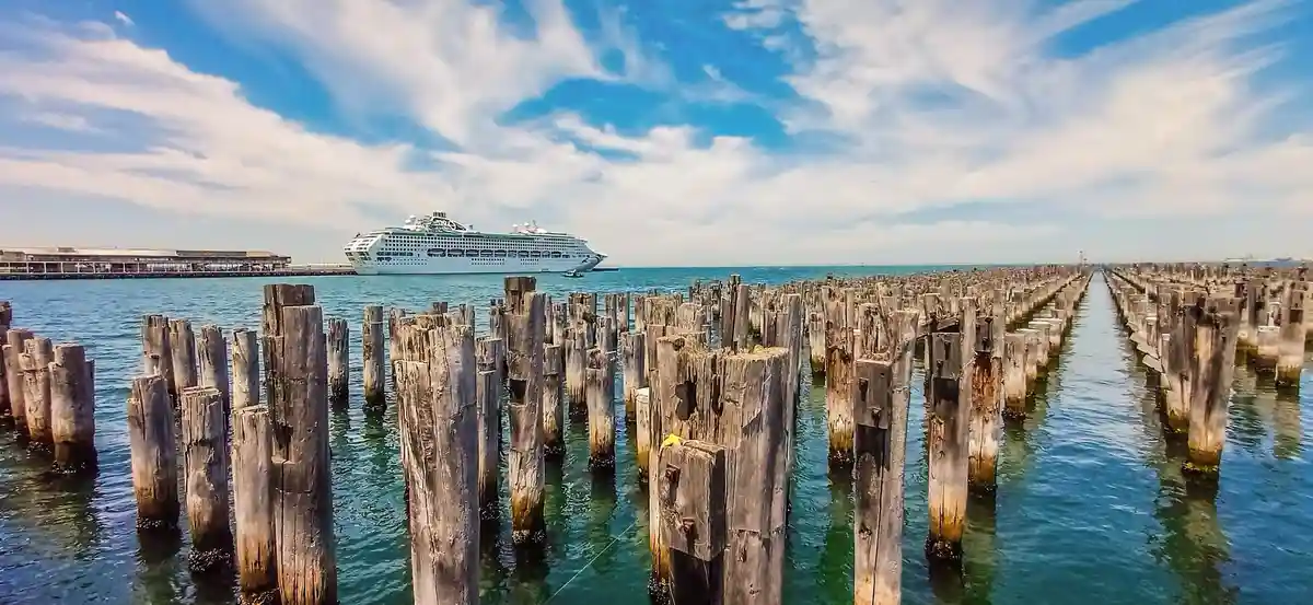 A wide-angle photo shows a large white cruise ship docked in the distance under a blue sky with wispy clouds. In the…