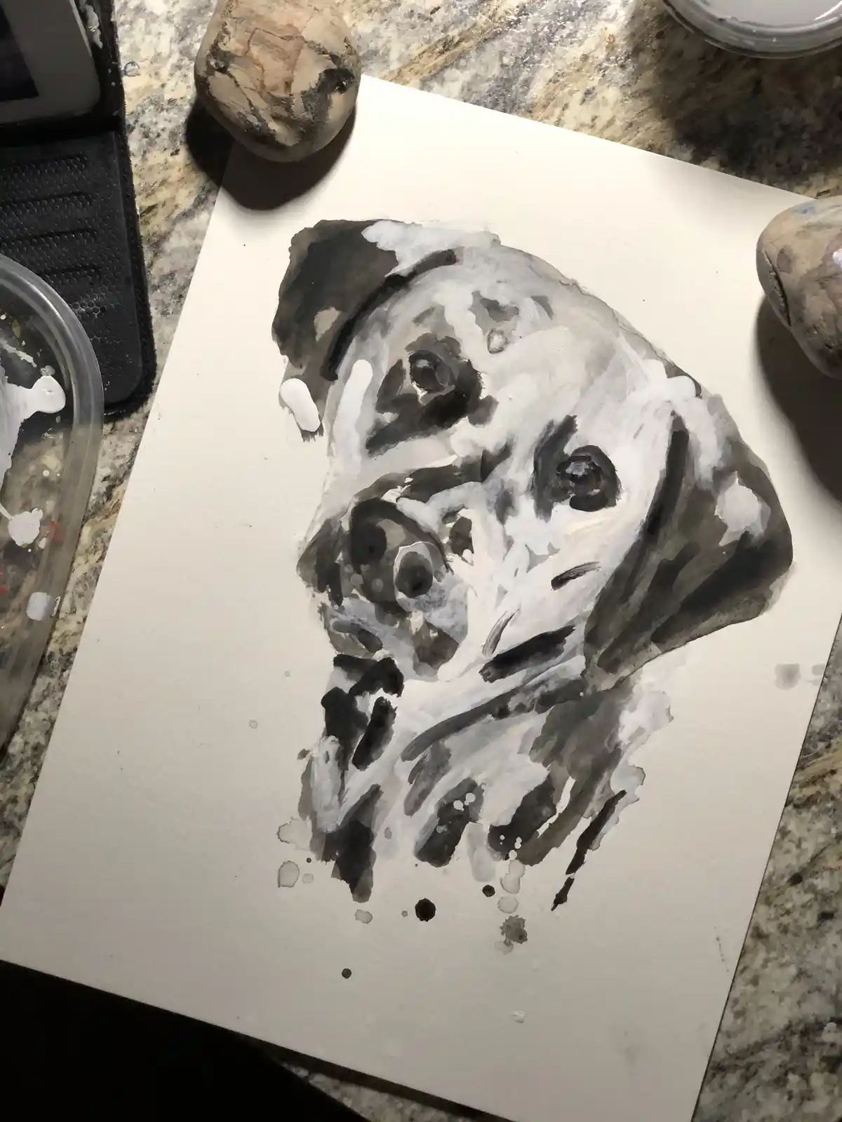 A watercolor painting on white paper depicts a dog's head with black and white splatters and smudges, looking somewhat…