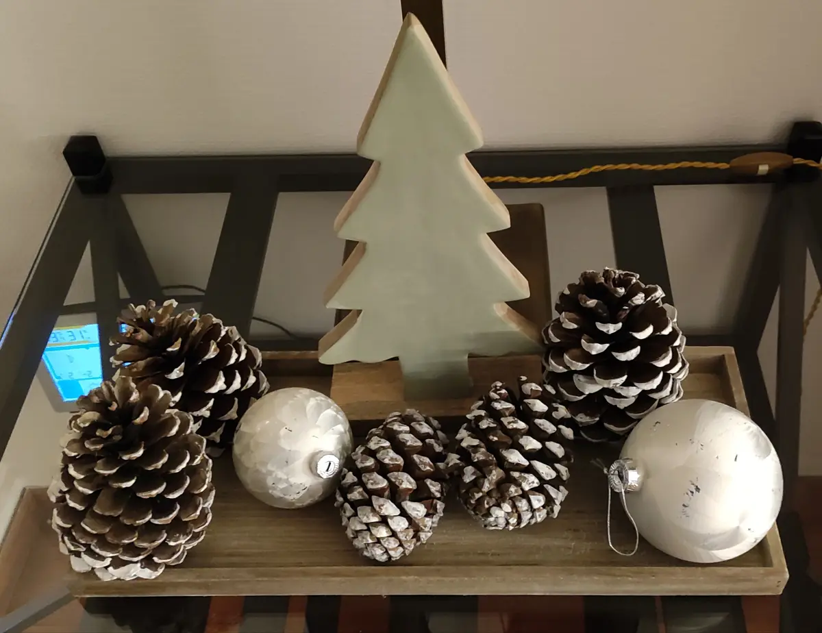 A wooden tray holds Christmas decorations including pinecones, white ornaments, and a white wooden tree. The pinecones are…