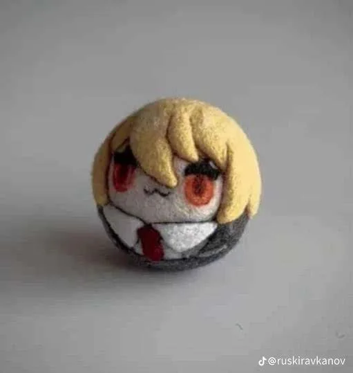 A small, round, felt craft depicts a stylized anime character with blonde hair, red eyes, and a white shirt with a red tie…