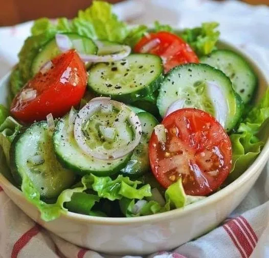 A close-up photo of a fresh salad in a white bowl, featuring sliced cucumbers, halved cherry tomatoes, thinly sliced red…