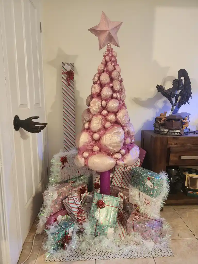 A pink, glittery Christmas tree made of stacked, irregularly shaped rocks sits surrounded by numerous wrapped presents on a…