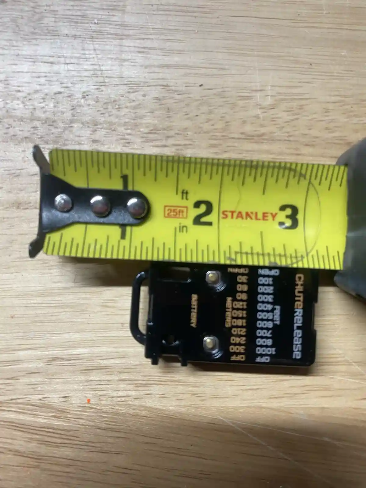A yellow Stanley 25ft tape measure is laid over a black Chuterelase remote control, which has a scale labeled 'Feet' and…