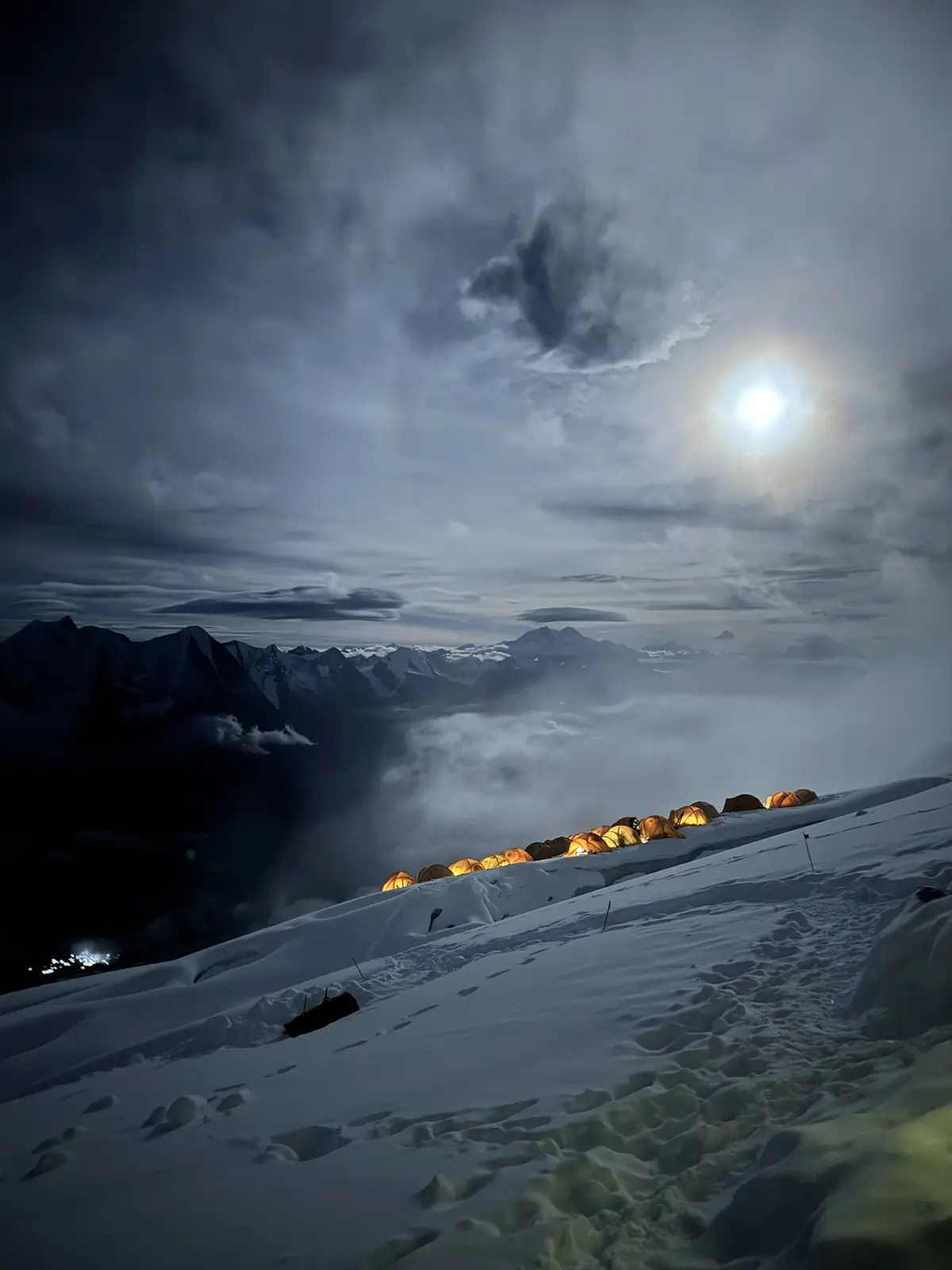 A row of illuminated orange tents is pitched on a snowy mountain ridge under a bright moon and cloudy sky, with distant…