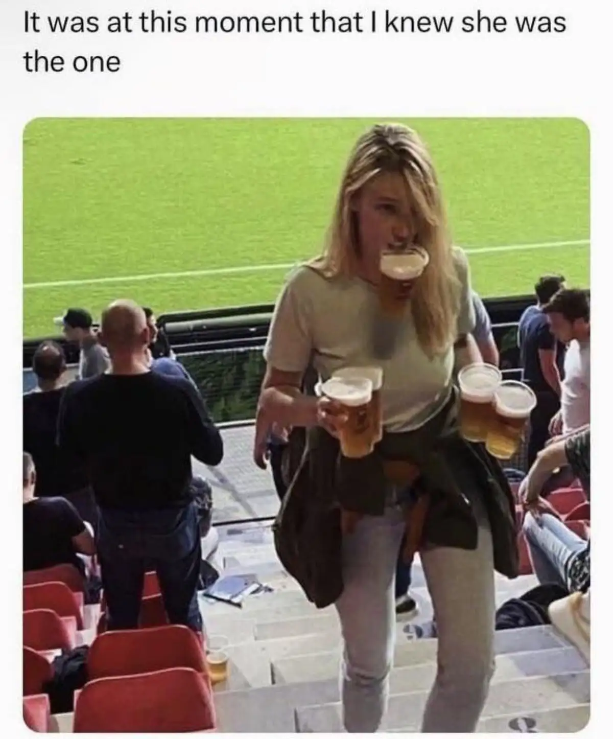 A woman with blonde hair, wearing a light-colored t-shirt and jeans, is walking down stadium stairs while holding multiple…